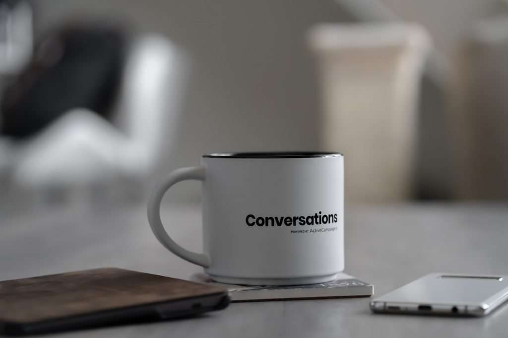 A white coffee mug with the word "Conversations" printed on it, accompanied by a smartphone and a wallet on a table, with a soft-focus background.