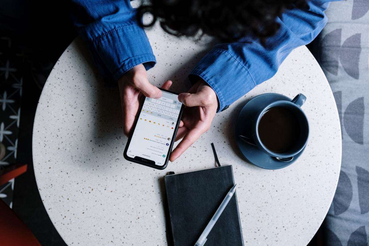 A person typing on a smartphone while seated at a table with a cup of coffee and a notebook.