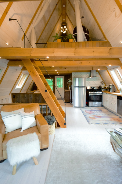 Interior view of a cozy A-frame cabin featuring wooden beams, a loft, and modern kitchen appliances, with a comfortable seating area and warm lighting.
