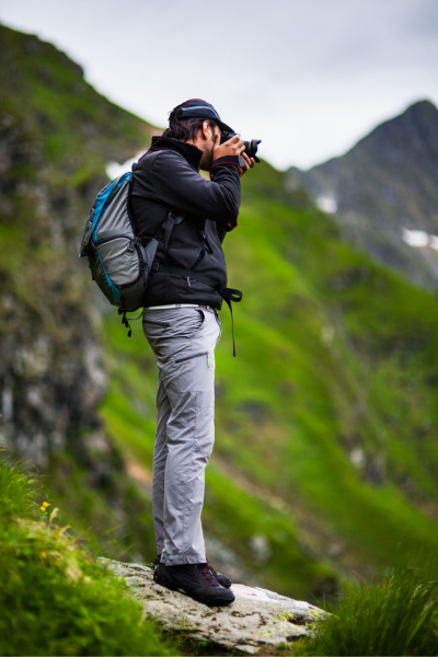 A hiker with a camera stands on a rocky ledge, surrounded by lush green mountains, as he focuses on capturing the stunning landscape.